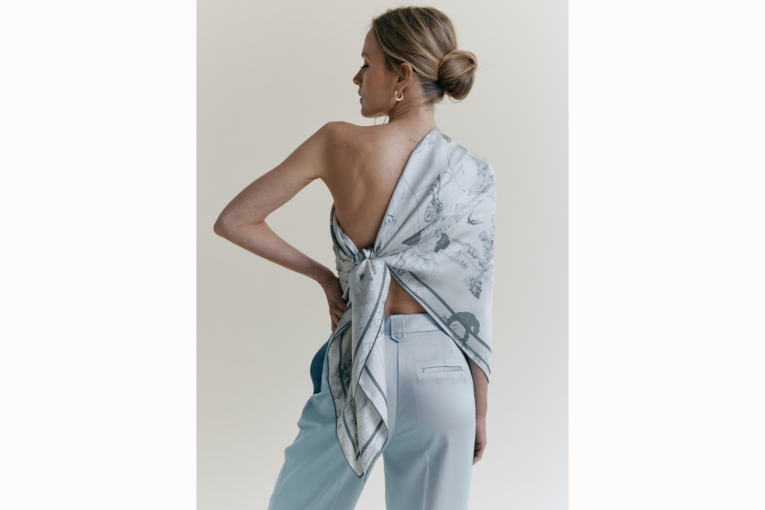 Female model faces her back to the camera wearing the pewter scarf styled as a one-shoulder top, tied on the back left side. She wears gold hoops and light blue pants. Her hair is styled up in a bun to highlight details of the scarf. 