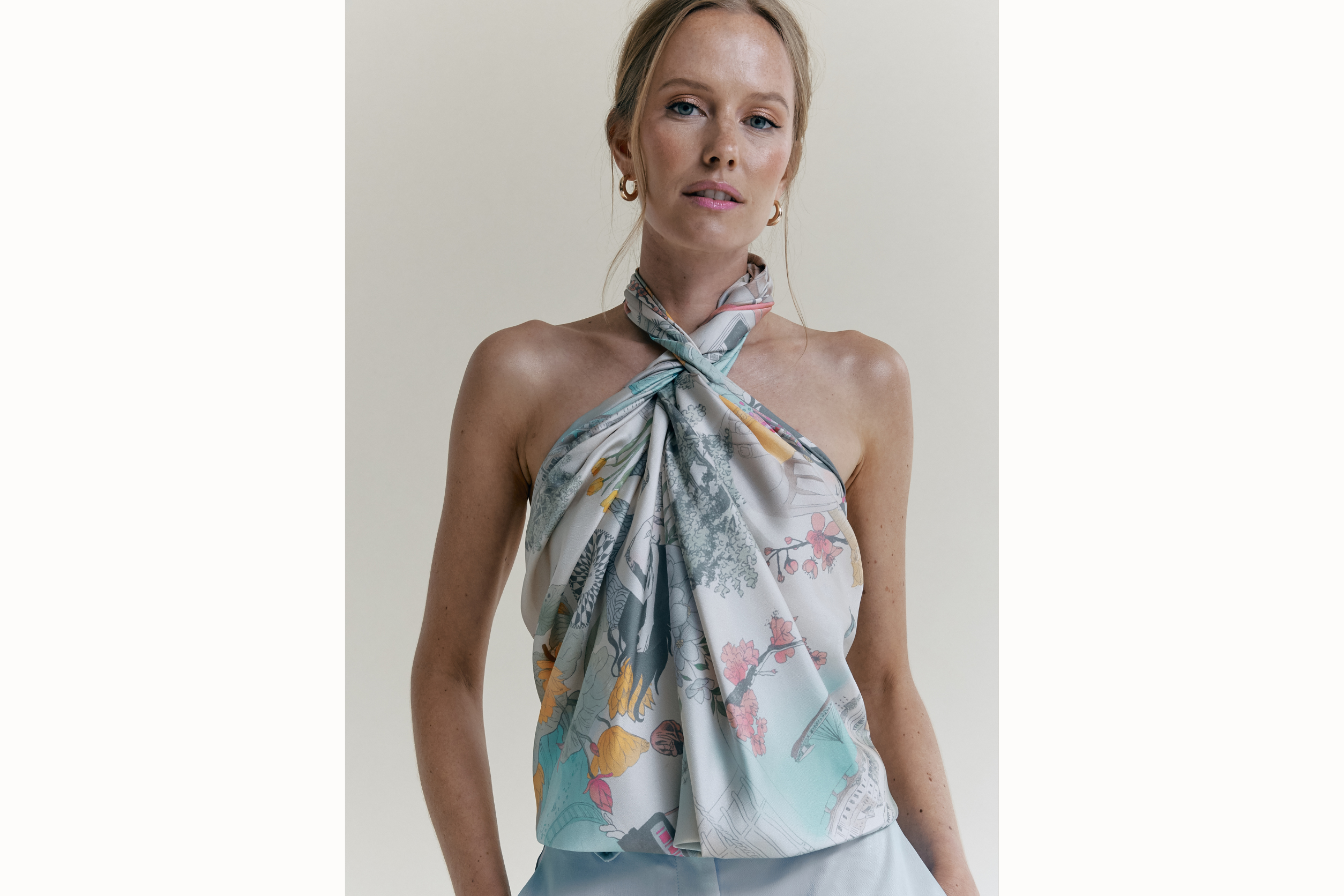Model wears watercolor version of the scarf styled as a halter top. Her hair is in a bun and she wears light blue pants. 