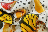 Close-up image of 100% silk square scarf featuring a motif of large-scale illustrated monarch wings and milkweed plants on a white background with imperfectly shaped black polka dots. Illustrates the lightly ridged texture of the silk twill along with the rich color tones and luminous nature of the silk scarf.