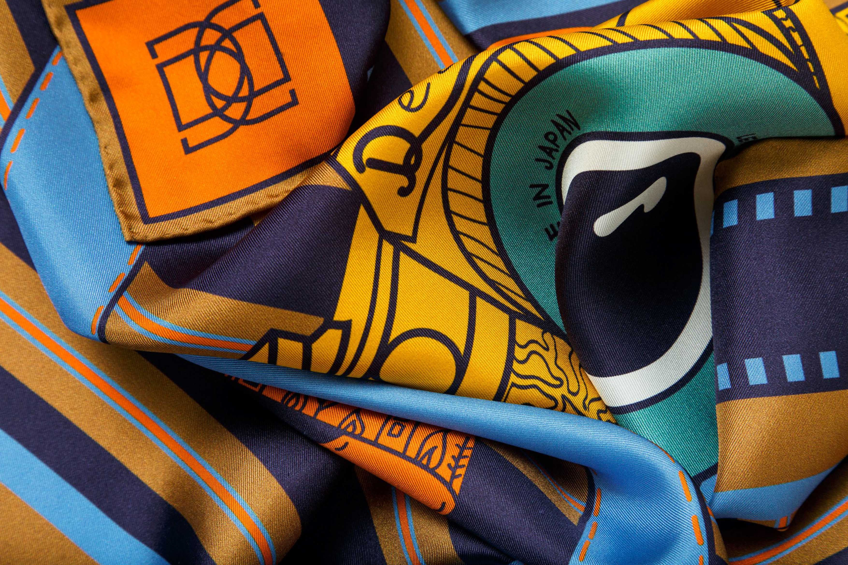 Rolled image of 100% silk square scarf featuring a motif of two large-scale illustrated film cameras and their straps splashed on top of a multi-color striped background. Blue film-inspired border around scarf. Design features colors such as orange, navy, camel, maize and a touch of aqua.