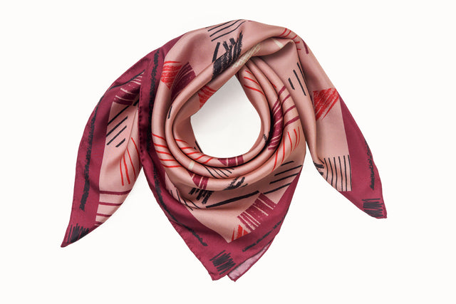 Rolled image of 100% silk square scarf featuring a motif of charcoal, cherry, raspberry and cream colored imperfect lines making geometric rectangle and triangle shapes throughout the scarf. Blush pink background and rich berry colored border on the scarf. Illustrates the lightly ridged texture of the silk twill along with the rich color tones and luminous nature of the silk scarf.