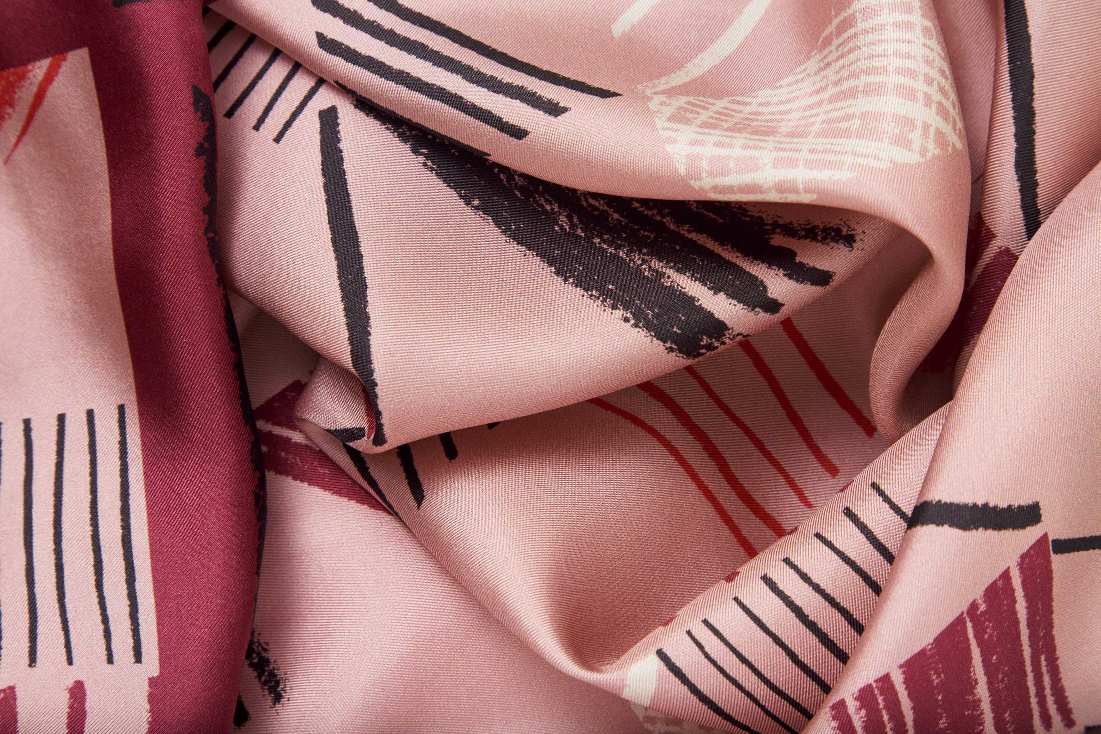 Close-up image of 100% silk square scarf featuring a motif of charcoal, cherry, raspberry and cream colored imperfect lines making geometric rectangle and triangle shapes throughout the scarf. Blush pink background and rich berry colored border on the scarf. Illustrates the lightly ridged texture of the silk twill along with the rich color tones and luminous nature of the silk scarf.