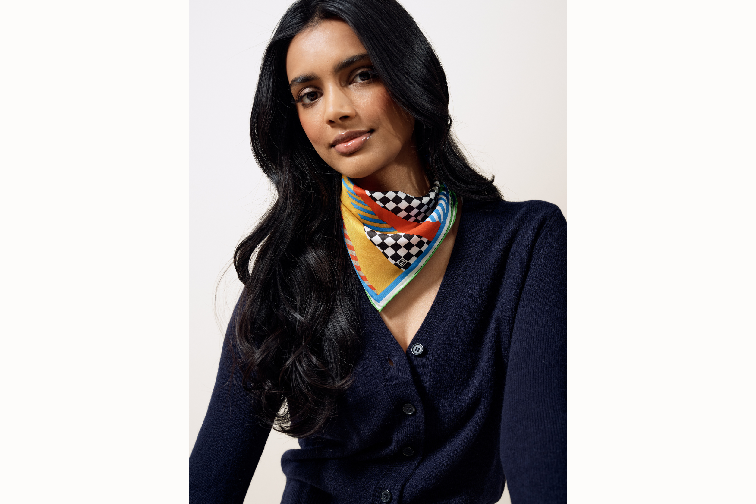 Image of female model wearing the scarf tied as a bandana with scarf draping over front of the base of her neck. Model also wearing a blue v-neck cardigan. .