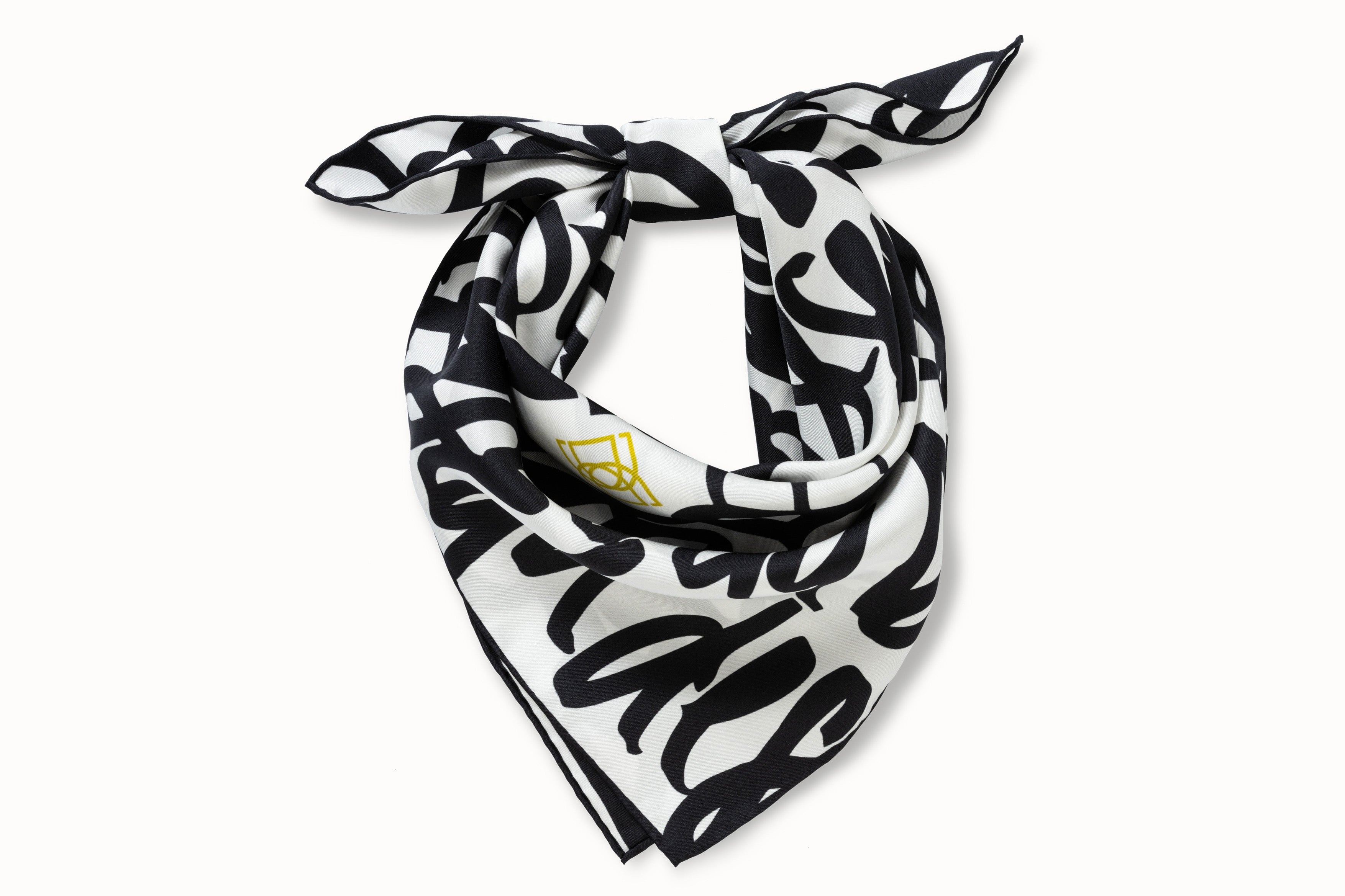 Rolled image of 100% silk square scarf featuring DESEDA written in black script on a repeat throughout the scarf on a white background. Features one small citron-colored DESEDA logo placed off-center in the design. Thin black border around the scarf’s edges.