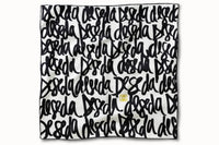 Flatlay image of 100% silk square scarf featuring DESEDA written in black script on a repeat throughout the scarf on a white background. Features one small citron-colored DESEDA logo placed off-center in the design. Thin black border around the scarf’s edges.