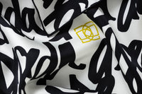 Close-up image of 100% silk square scarf featuring DESEDA written in black script on a repeat throughout the scarf on a white background. Features one small citron-colored DESEDA logo placed off-center in the design. Thin black border around the scarf’s edges. Illustrates the lightly ridged texture of the silk twill along with the rich color tones and luminous nature of the silk scarf.