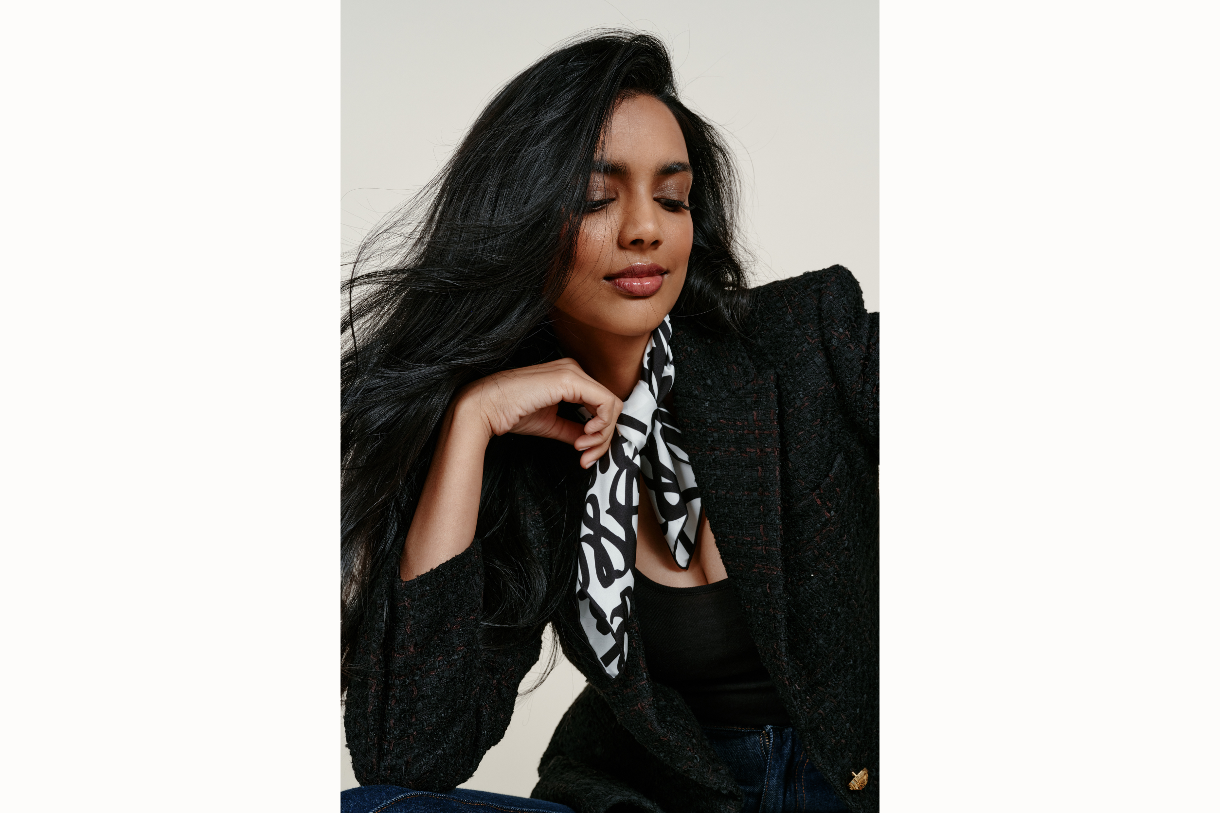 Image of a female model leaning in but looking away from the shot, wearing the black and white scarf rolled around and tied at the neck. Model sports a black blazer while touching the scarf with her right hand. 