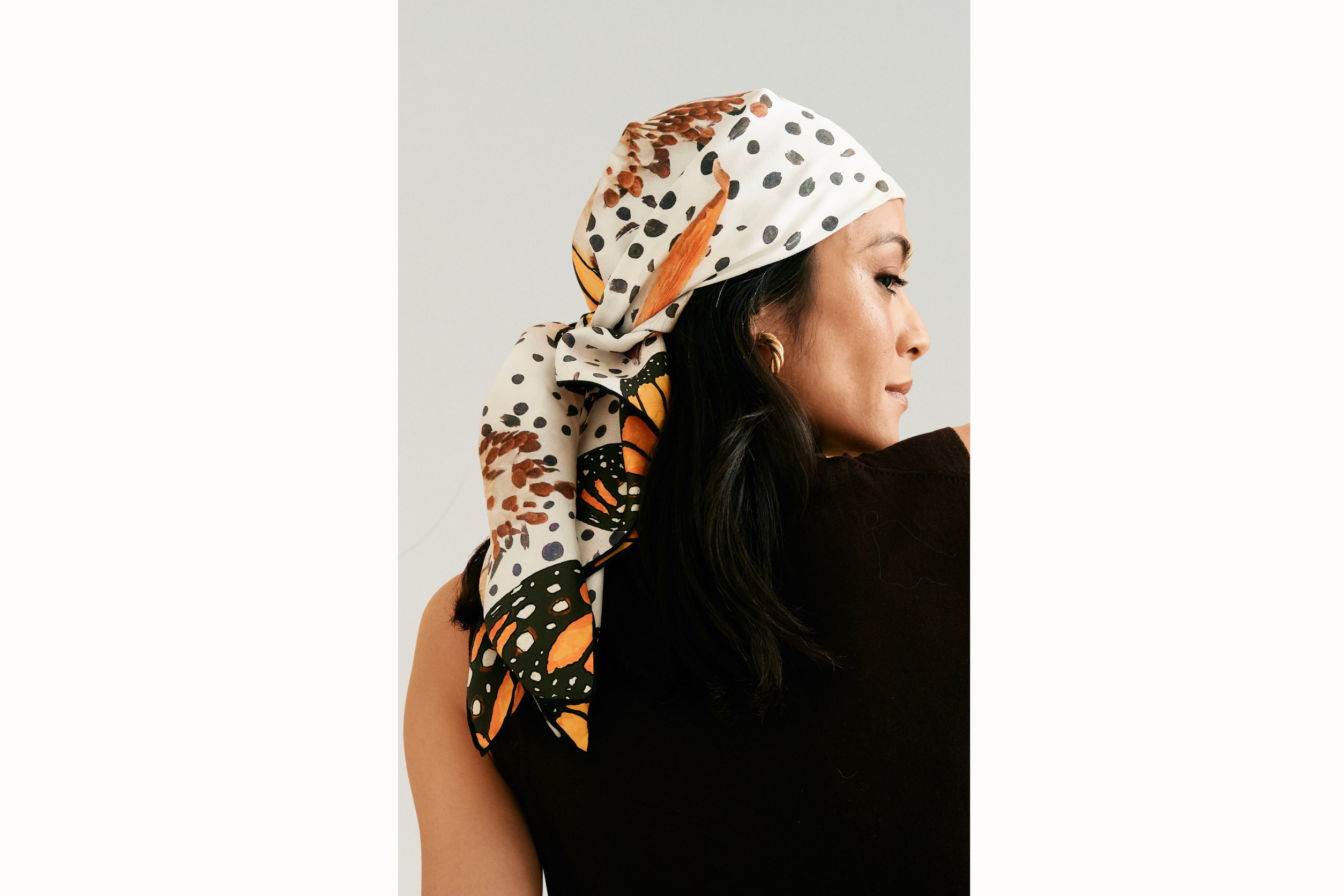Female model wears scarf as a headscarf, knotted in the back. She wears gold hoops and a black tank as she looks over her shoulder.
