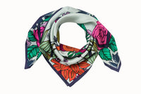 Rolled image of 100% silk square scarf featuring a motif of bright, bold florals and greenery around the border as well as in the center of the scarf wrapped around two knotted garden snakes. Colors featured in include various shades of vibrant green, orange, lilac and rose pink on a very light and soft mint background.