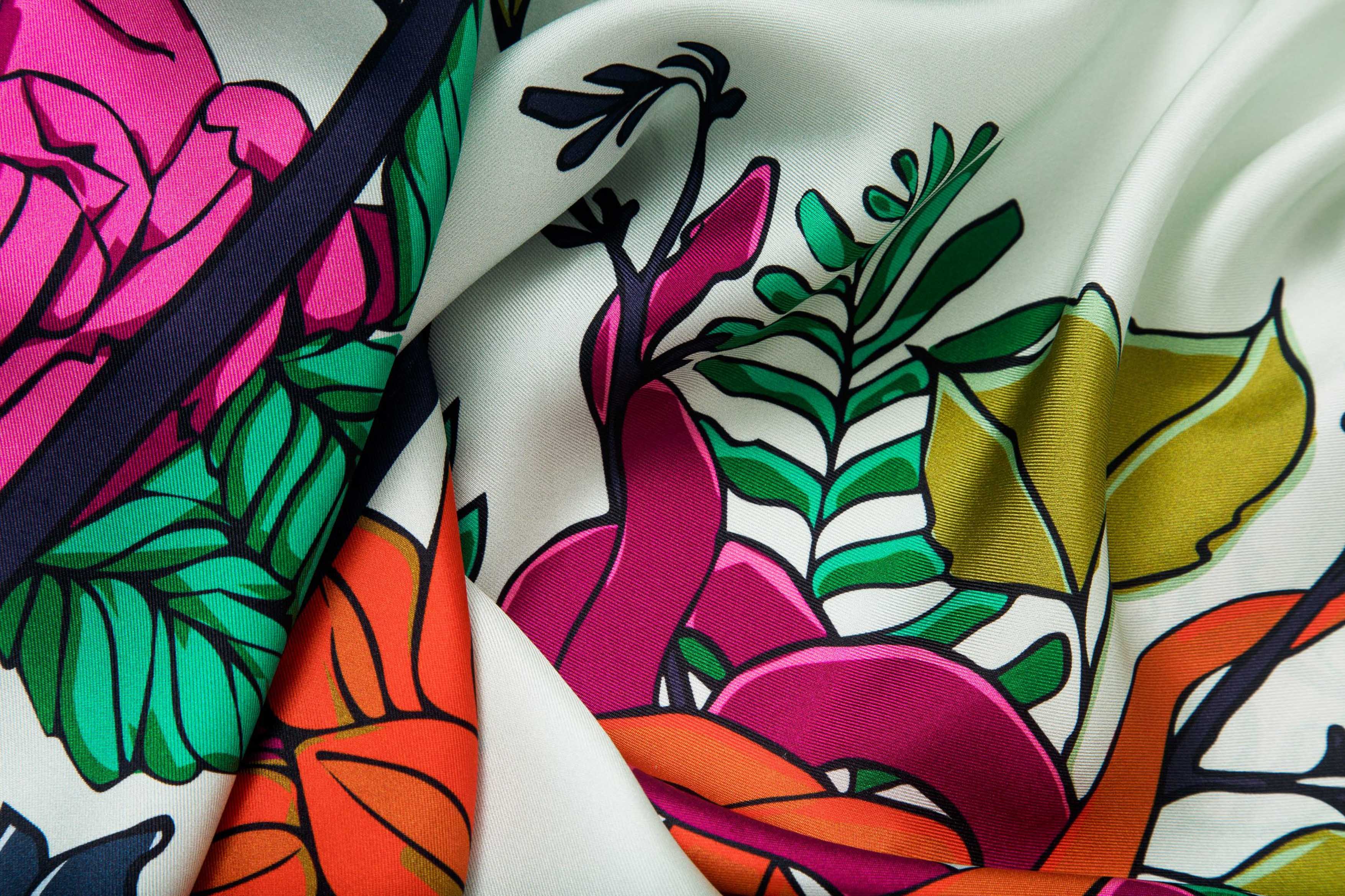 Close-up image of 100% silk square scarf featuring a motif of bright, bold florals and greenery around the border as well as in the center of the scarf wrapped around two knotted garden snakes. Colors featured in include various shades of vibrant green, orange, lilac and rose pink on a very light and soft mint background. Illustrates the lightly ridged texture of the silk twill along with the rich color tones and luminous nature of the silk scarf.
