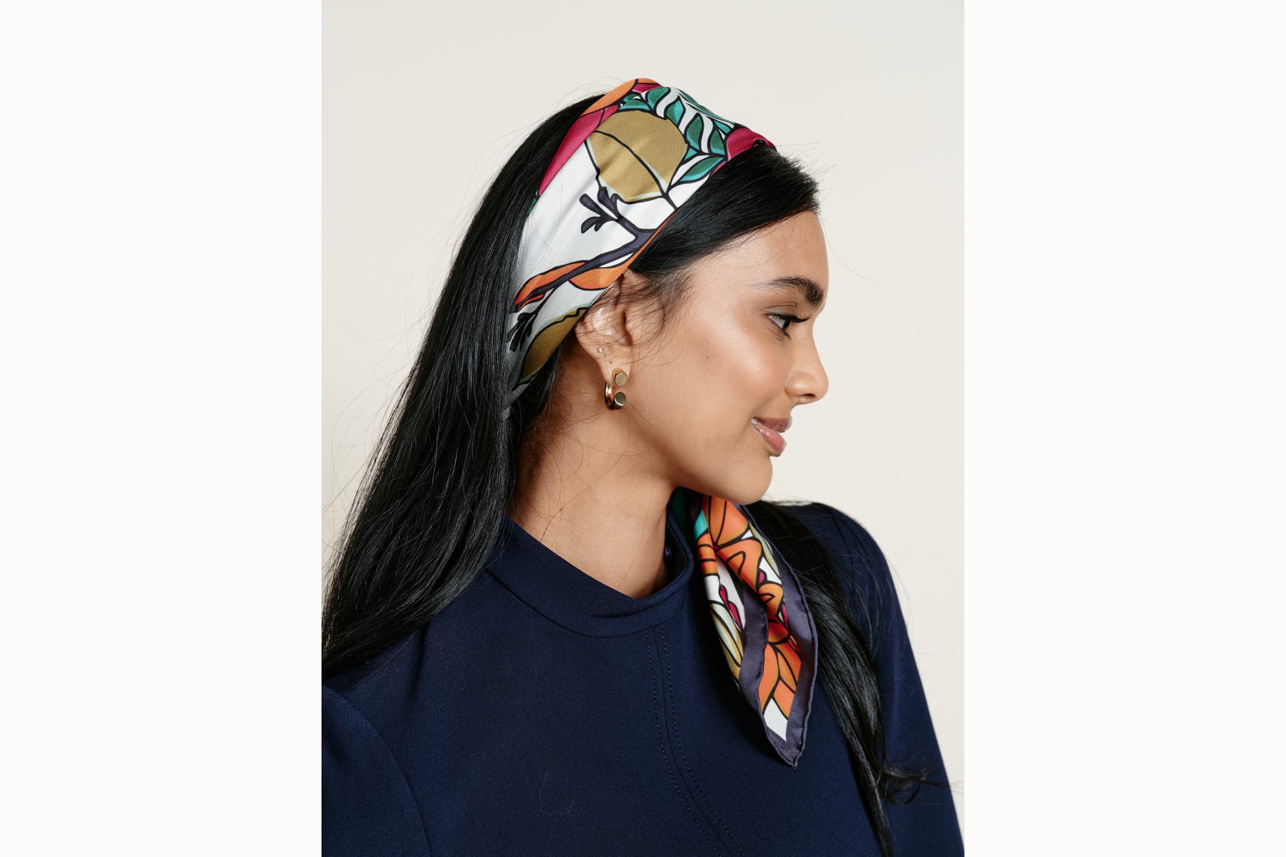 Female model wearing scarf folded and tied as a headband with the ends on the scarf pulled over her shoulder. She looks down and away from the camera, slightly turned and is also wearing a navy mock neck. 