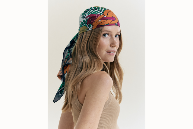 Female model wearing scarf styled as a headscarf and tied at the back of her head. She wears a neutral colored tank and looks over her shoulder into the camera. . 