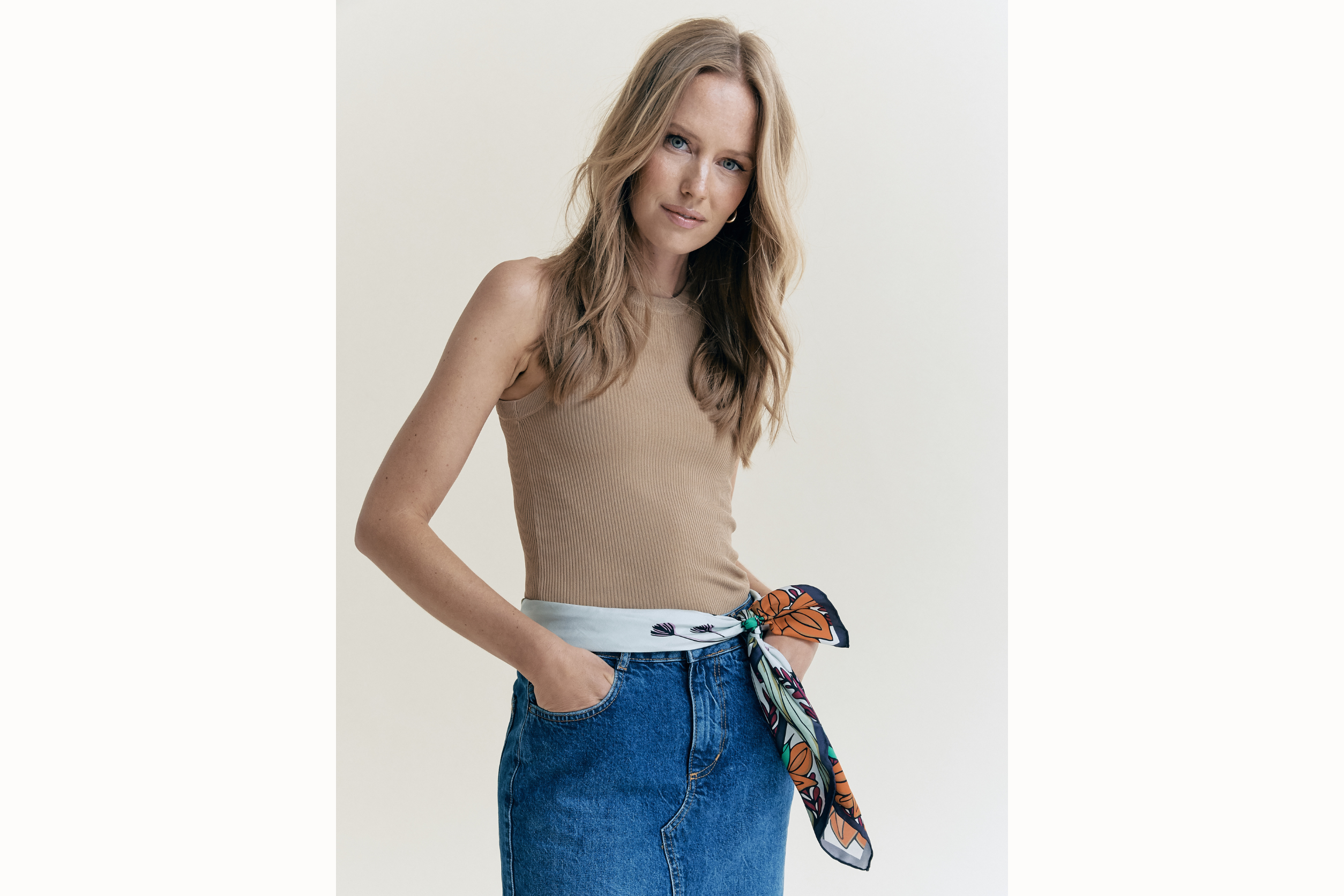 model wears scarf folded and tied around her waist as a belt. She has her hands in the pockets of her denim skirt and also sports a brown tank top. 