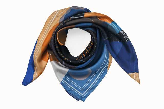 Rolled image of 100% silk square scarf featuring a cosmic collage motif, including a large planet in the center surrounded by the moon phases and stars in outer space. Colors used include shades of warm oranges and blues as well as white and black details.