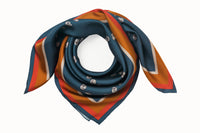 Rolled image of 100% silk square scarf featuring a motif of cream embroidery knots arranged to represent the DESEDA logo on a steely blue background. Features a multi-stripe border in shades of pumpkin, cream, and coral.