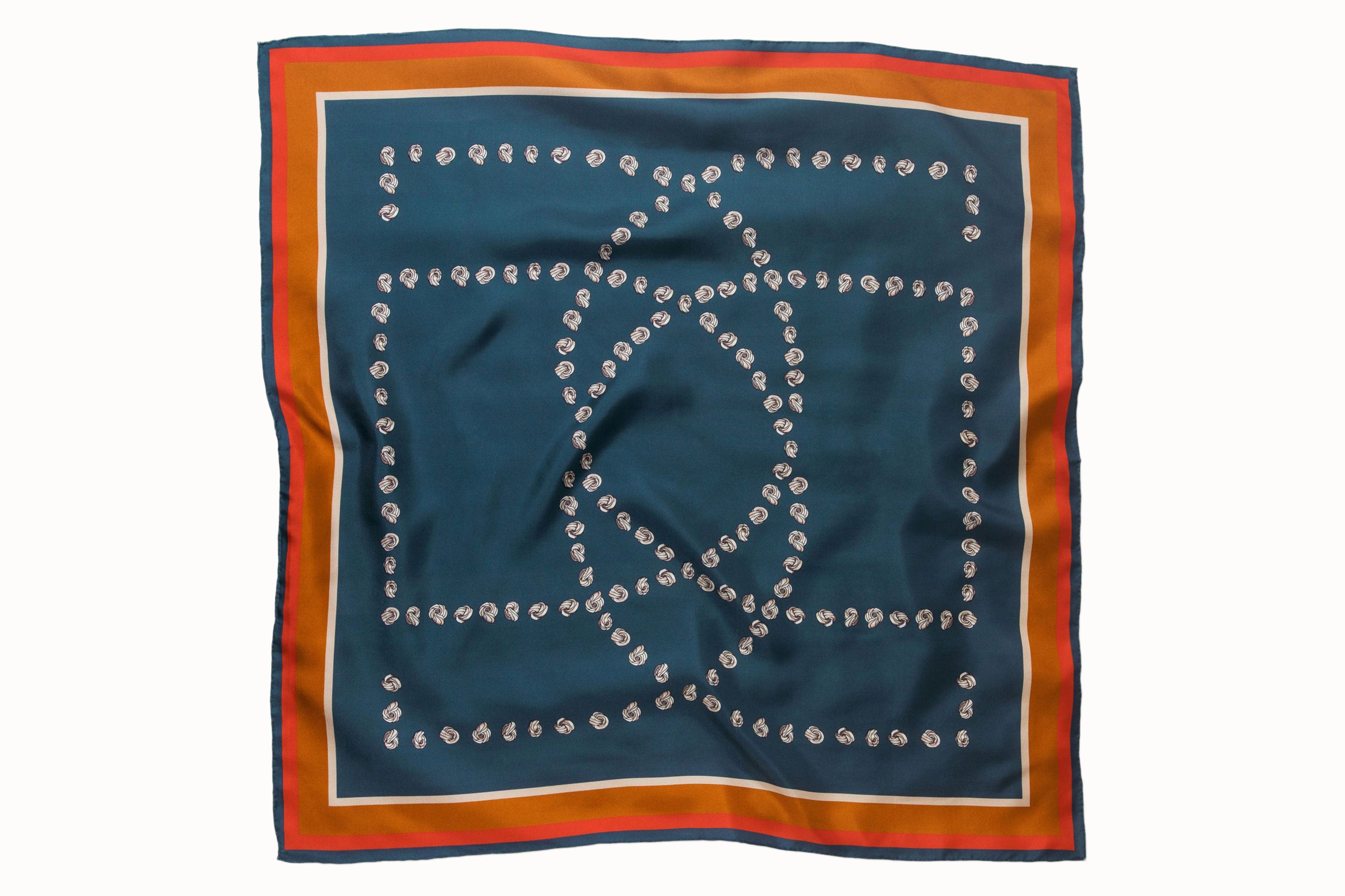 Flatlay image of 100% silk square scarf featuring a motif of cream embroidery knots arranged to represent the DESEDA logo on a steely blue background. Features a multi-stripe border in shades of pumpkin, cream, and coral.