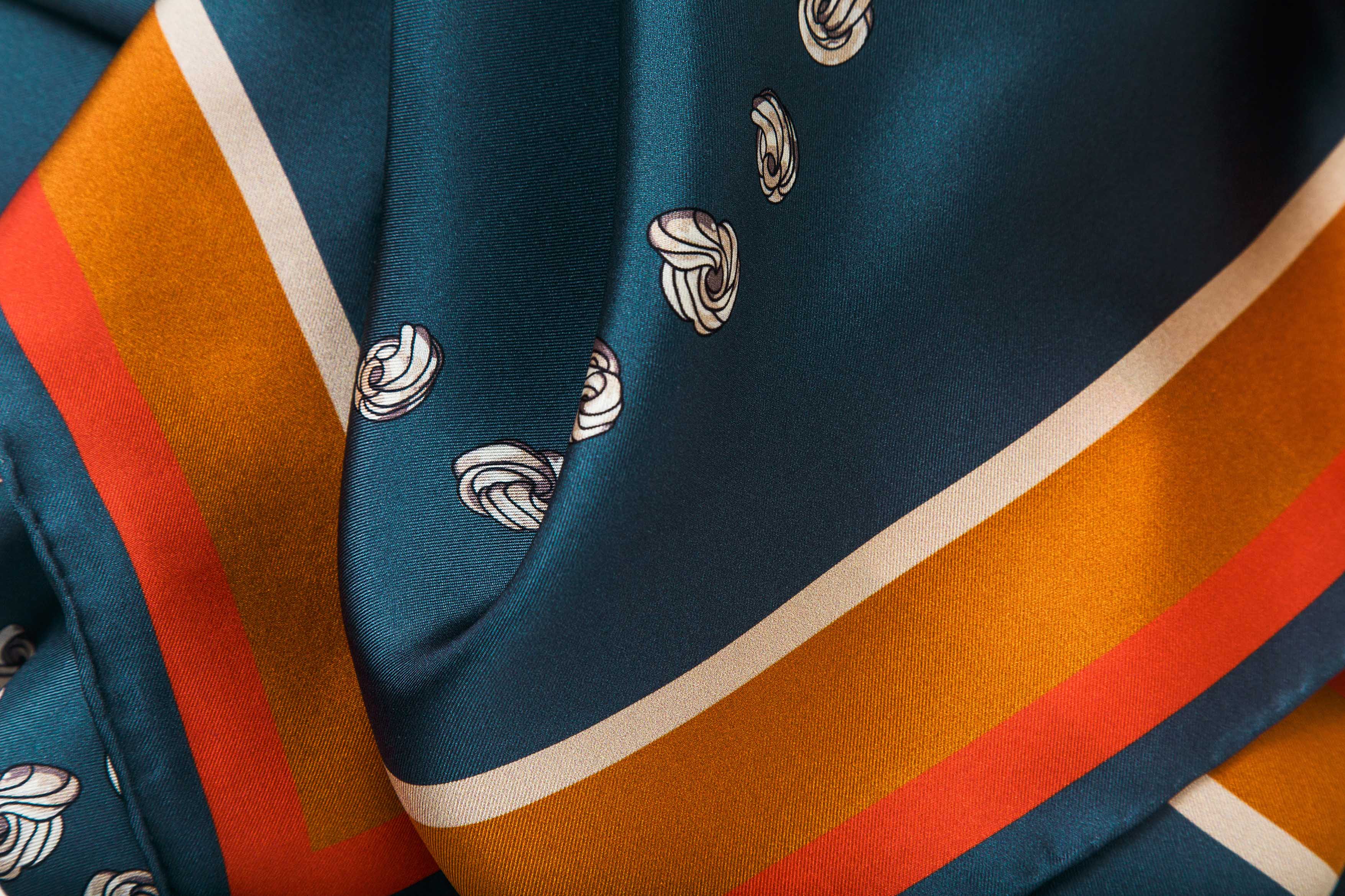 Close-up image of 100% silk square scarf featuring a motif of cream embroidery knots arranged to represent the DESEDA logo on a steely blue background. Features a multi-stripe border in shades of pumpkin, cream, and coral. Illustrates the lightly ridged texture of the silk twill along with the rich color tones and luminous nature of the silk scarf.