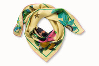 Rolled image of 100% silk square scarf featuring two painted two mermaids resting on large sea shells, a pink flower and green seaweed on a lemon yellow background with small butterfly accents. Thin green border around the edges of the scarf, layered with green leaves, seashells and lemons.