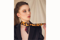 Image of female model wearing the scarf tied around the base of her neck and secured with a simple knot. The tail ends of the scarf drape down her chest. Model is also wearing a black blazer.