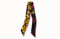Flatlay image of 100% silk ribbon style scarf, 2” wide by 32” long. Features an abstract leopard print design in shades of black, brown and yellow on a soft orange-colored background. The opposite side of the scarf features a series of bursting abstract floral images in shades of pink, orange and yellow on a black background.
